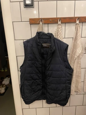 Svart dunväst från Ocean Pacific - Snygg svart dunväst från Ocean Pacific med quiltat mönster och hög krage. Västen har dragkedja framtill, två sidofickor och en innerficka med knapp. Perfekt för lager på lager och enkel att matcha till olika outfits.