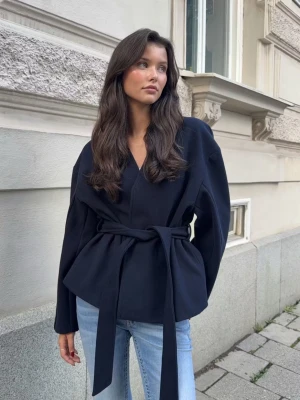 Svart kappa med knytskärp - Stilren svart kavaj med avslappnad passform och brett knytskärp i midjan. Kavajen har V-ringning, långa ärmar och är tillverkad i ett mjukt tyg som ger en modern siluett. Perfekt att styla med jeans för en trendig look.