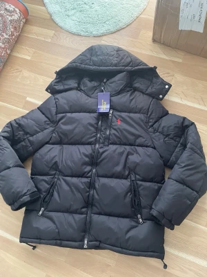 Polo Ralph Lauren Pufferjacket - Svart pufferjacka från Polo Ralph Lauren med avtagbar huva, två stora dragkedjefickor och tydlig röd broderad logga på bröstet. Jackan har breda kanaler och grov dragkedja framtill. Materialet är glansigt och tåligt, perfekt för kalla vinterdagar.