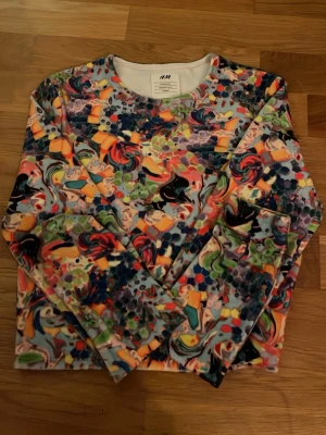 Färgglad mönstrad tröja från H&M - Säljer en långärmad tröja från H&M med ett färgstarkt abstrakt mönster i orange, blått, grönt, svart och lila. Tröjan har rund halsringning och är tillverkad i mjukt material, perfekt för dig som gillar statement-plagg.
