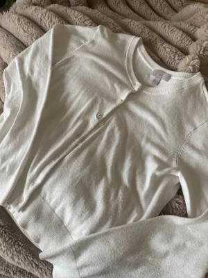 Vit kofta från H&M med knappar - Klassisk vit kofta från H&M med rund halsringning och knäppning framtill. Tillverkad i mjukt, tunt material med ribbade muddar vid ärmslut och nederkant. Perfekt att slänga över en topp eller klänning för en clean look.