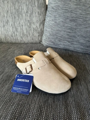 Beige Birkenstock Boston tofflor - Säljer ett par snygga beige Birkenstock Boston tofflor i mocka med justerbart spänne i metall. Klassisk slip-in modell med bekväm fotbädd och svart sula. Perfekta för dig som gillar stilren och enkel design.