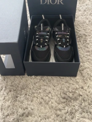 Svarta Dior B22 sneakers - Säljer ett par svarta Dior B22 sneakers med chunky sula och meshdetaljer. Skorna har inslag av iriserande färger och vita detaljer på snörningen. Märket syns tydligt på sidan och tungan. Perfekta för dig som vill ha en statement sneaker.