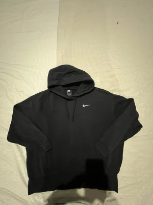 Svart Nike hoodie med logga - Klassisk svart hoodie från Nike med vit broderad Swoosh-logga på bröstet. Hoodien har huva med dragsko och ribbade muddar vid ärmar och nederkant. Tillverkad i mjukt bomullsmaterial som är skönt att bära.