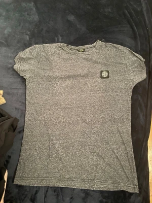 Gråmelerad Stone Island t-shirt - Säljer en gråmelerad t-shirt från Stone Island med klassisk patch på bröstet. T-shirten har rund hals och korta ärmar, perfekt för en clean och avslappnad stil. Materialet känns mjukt och skönt mot huden.