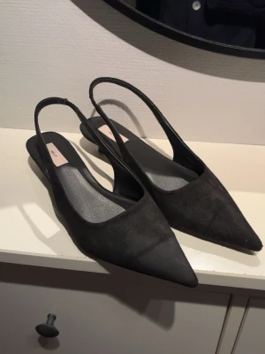 Svarta spetsiga pumps - Säljer ett par svarta slingback pumps med spetsig tå och öppen häl. Skorna har en låg klack och är tillverkade i mockaimitation som ger en snygg och stilren look. De är fina utanpå men har lite lim från plåster inuti skon som inte går bort men det är inget som påverkar funktionen. Säljer då de var lite för små för mig😊