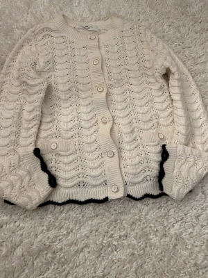 Vit virkad kofta med vågmönster - Säljer en söt vit kofta med vågigt virkat mönster och svarta detaljer längst ner. Koftan har rund hals, knappar framtill och ribbade muddar. 