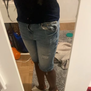Ljusblå jeansshorts med slitningar - Säljer ett par ljusblå jeansshorts med slitna detaljer och klassisk femficksdesign. Shortsen har normal passform och går ner till knäna. Perfekta för sommaren och enkla att matcha med olika toppar.