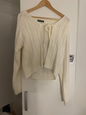 Vit kabelstickad kofta från Shein - Säljer en vit kabelstickad kofta från Shein med knytband framtill. Koftan har lång ärm, ribbade muddar och ett snyggt kabelmönster över hela plagget. Perfekt att slänga över en topp för en chill look.