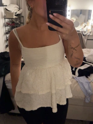 Vit volangtop med smala axelband - Supersöt vit topp med volanger och smala axelband. Toppen har en lätt rynkad struktur och är perfekt för varma dagar. Den är ärmlös och har en luftig passform som känns både trendig och bekväm.