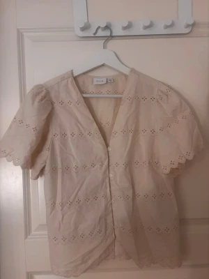 Beige blus med broderi från Vila - Söt beige blus från Vila med korta puffärmar och v-ringning. Blusen har dekorativa broderade hål och knäppning framtill. Tillverkad i ett lätt bomullstyg med vågig kant vid ärmslut och nederkant. Blusen är använd vi få tal gånger. Pris kan diskuteras 