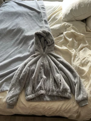 Grå teddyjacka med huva från Bershka - Supermjuk och fluffig teddyjacka i ljusgrått från Bershka. Jackan har dragkedja framtill, stor huva och breda muddar vid ärmsluten. Perfekt för kalla dagar när du vill vara både varm och stylish.