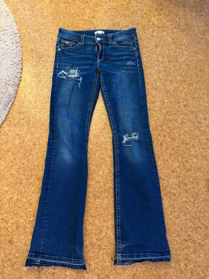 Blå slitna bootcut jeans - Säljer ett par blå bootcut jeans med slitningar och hål på låret och knät. Jeansen har råa kanter nertill och klassisk femficksmodell. Perfekta för en avslappnad och trendig look. Materialet är denim i bomull.