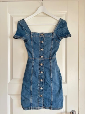 Kort jeansklänning med knappar - Snygg kort jeansklänning med markerad midja och knappar framtill. Klänningen har korta ärmar, fyrkantig urringning. Bra passform ! Klänningen kan ha tecken på användning 