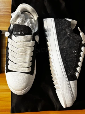 Svarta och vita sneakers från Numeris - Snygga sneakers från Numeris i svart denimtyg med vita detaljer och breda vita skosnören. Skorna har en chunky vit sula med coolt sågmönster på sidan och vit hälkappa i strukturmaterial. Perfekta för dig som vill sticka ut med din stil.