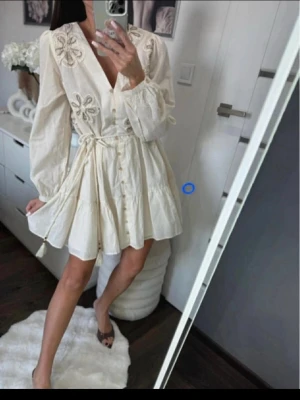  Ny slutsåld Krämvit kortklänning med broderi - Söt krämvit kortklänning med broderade blommor på axlarna, v-ringad hals och knappar framtill. Klänningen har långa puffärmar och ett knytband i midjan för en feminin siluett. Perfekt för sommarens alla tillfällen.