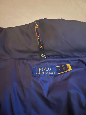 Blå pufferjacka från Polo Ralph Lauren - Snygg blå pufferjacka från Polo Ralph Lauren med klassisk design. Jackan har en hög krage och insida med märkets logotyp. Perfekt för kalla dagar och ger en stilren look med sin djupblå färg och quiltade material. Passar också bra med många outfits.         Pris kan sänkas 