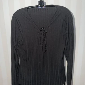 Svart ribbad topp med snörning - Snygg svart ribbad topp med lång ärm och snörning framtill i urringningen. Toppen har en v-ringad form och är tillverkad i ett stretchigt material som sitter bekvämt på kroppen. Perfekt att styla med jeans eller kjol för en trendig look.