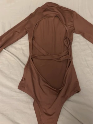 Brun långärmad body med cut-outs - Snygg brun body med långa ärmar och stora cut-outs på ryggen. Tillverkad i ett stretchigt, glansigt material som sitter tight mot kroppen. Perfekt för dig som vill ha något trendigt och vågat. Inga synliga märken eller loggor.