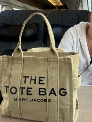  tote bag oliv grön - En olivgrön tote bag från Marc Jacobs med tydlig svart logga på framsidan. Väskan är i canvasmaterial och har två handtag. Perfekt storlek för att bära med sig det viktigaste och har en stilren desigin. Väldigt rymlig och perfekt söt storlek. Medföljer en bred rem för att bära över sig eller hängandes på axeln. Pris kan diskuteras. 
