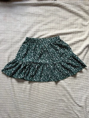 Grön blommig volangkjol - Supersöt grön kortkjol med vitt blommönster och volangkant. Kjolen har resår i midjan för skön passform och är perfekt för vår och sommar. Tillverkad i ett lätt och luftigt material som känns bekvämt hela dagen.