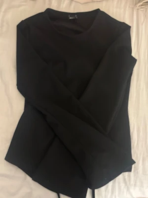 Svart långärmad topp med knyt - Stilren svart långärmad topp med rund hals och knyt i midjan som ger en snygg omlott-effekt baktill. Perfekt att matcha med både jeans och kjol för en trendig look. Materialet känns mjukt och stretchigt.