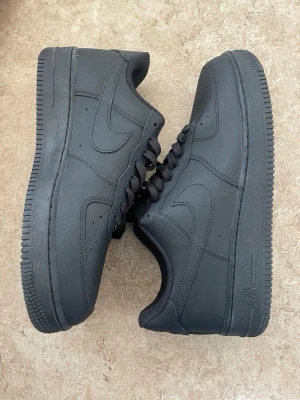 Svarta Nike Air Force 1 sneakers - Klassiska helsvarta Nike Air Force 1 sneakers i lågt utförande. Skorna har en stilren design med diskret Nike Swoosh på sidorna, perforeringar på tån för ventilation och robust gummisula. Tillverkade i slätt läder med matchande svarta skosnören.