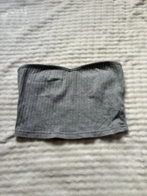 Grå ribbad bandeau tubtopp - Säljer en enkel och stilren grå bandeau tubtopp med ribbad struktur. Toppen är ärmlös och har en mjuk, stretchig passform som sitter snyggt på kroppen. Perfekt att matcha med höga jeans eller kjol för en clean look.