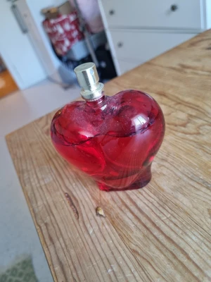 Röd hjärtformad parfymflaska 100ml - Säljer en Eau de Parfum i en snygg hjärtformad flaska i rött glas. Flaskan rymmer 100 ml och har en silverfärgad spraykork. Perfekt för dig som vill ha en unik och dekorativ parfymflaska på hyllan.