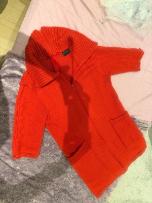 Orang stickad kofta med dragkedja betti Kai rand - Snygg orange stickad kofta med dragkedja bak och dragkedjor på ärmarna också, två stora fickor, korta ärmar och en bred, ribbad krage som ger en cool look. Perfekt att slänga över en t-shirt för extra värme och stil, från märket Betti Kai Rand.