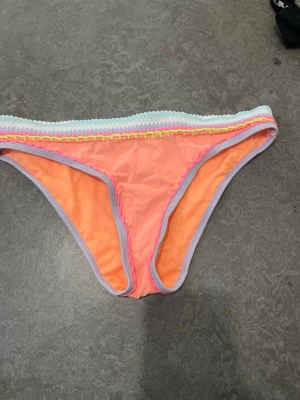 Neonorange bikiniunderdel med pastellkant - Säljer en snygg bikiniunderdel i neonorange med pastellfärgade kanter i ljuslila, mintgrönt och gult. Modellen har låg midja och färgglada detaljer upptill som ger en fräsch och ungdomlig look. Perfekt för strandhäng och pooldagar!