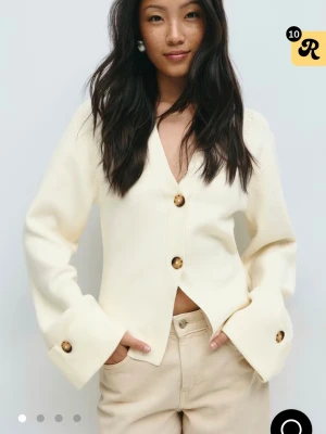 Krämvit kofta med stora knappar - Stilren krämvit kofta med v-ringning och stora bruna knappar framtill och vid ärmsluten. Koftan har en rak passform, långa ärmar och är tillverkad i ett mjukt, tjockare material som ger en exklusiv känsla.