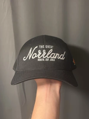 Svart keps The Great Norrland SQRTN - Svart keps från SQRTN med broderad text 'The Great Norrland' framtill och 'Sqrtn & Company' baktill. Justerbar snapback och böjd skärm. Klassisk streetstil med vit brodyr, perfekt för dig som gillar norrländsk vibe.