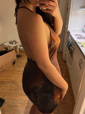 Tie dye kort klänning med öppen rygg - Säljer en kort klänning i tie dye-mönster med toner av orange, brun och svart. Klänningen är ärmlös, har smala axelband och en öppen rygg. Materialet känns mjukt och stretchigt, troligen polyester eller bomullsmix. Perfekt för dig som gillar en unik och trendig stil.