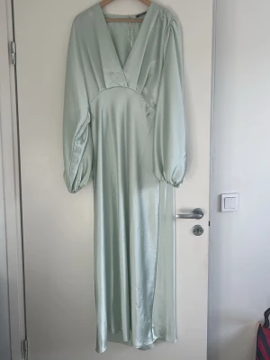 Ljusgrön långklänning med ballongärm - Elegant ljusgrön långklänning i satinliknande material med djup v-ringning och långa ballongärmar. Klänningen har en mjuk glans och en smickrande siluett som ger en lyxig känsla. Enbart använd en gång, kan skicka bilder på hur den sitter på om det önskas.