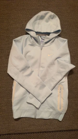 Ljusblå hoodie från Nike Nocta - Snygg ljusblå hoodie från Nike med dragkedja framtill och två fickor med dragkedja. Klassisk huva och diskret Nike-logga på bröstet. Tillverkad i mjukt material som känns skönt mot huden. Perfekt för en avslappnad och sportig stil. 