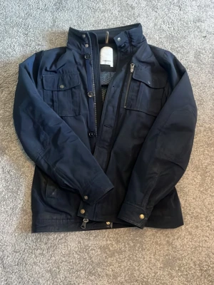 Mörkblå vindjacka med fickor - Snygg mörkblå vindjacka med flera praktiska fickor och dragkedja framtill. Jackan har hög krage, knappdetaljer och en clean, modern look. Perfekt för dig som vill ha en stilren jacka till vardags. Materialet är slitstarkt och jackan har lång ärm.