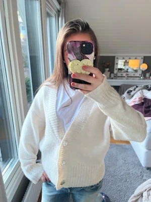 Vit  kofta med knappar - Mysig vit kofta med stora silvriga knappar och bred ribbad mudd vid ärmslut och nederkant. Koftan har en djup v-ringning och är gjord i ett mjukt, fluffigt material. Perfekt att slänga över en t-shirt för en chill look.