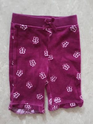 Byxor - Söta lila byxor med vita broderade fjärilar och volangkant nedtill. Byxan har en liten rosett i midjan och är tillverkade i mjuk bomull, perfekta för varma dagar.