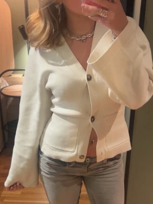 Vit ribbad kofta med knappar - Säljer en stilren vit kofta med bred ribbad midja och muddar. Koftan har djup V-ringning, stora silvriga knappar och två fickor framtill. Perfekt att matcha med jeans för en clean look.