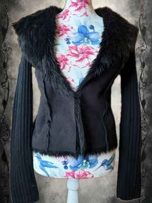 Svart cardigan med fuskpälsdetaljer - Stilren svart cardiganjacka med bred fuskpäls runt krage och öppning. Ärmarna är ribbstickade och ger en cool kontrast till den mjuka, släta kroppen. Jackan har en figurnära passform och är perfekt för dig som gillar edgy och mysig stil.