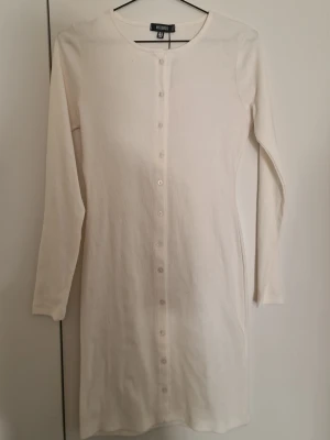 Vit långärmad klänning  - En enkel och stilren vit klänning med lång ärm och knappar hela vägen framtill. Klänningen har rak passform och är gjord i mjuk bomull, perfekt för en clean och avsl tort look.