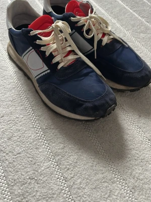 Philip model ”Runners”  - Snygga sneakers i blått och vitt med röda detaljer och vita skosnören. Ovandelen är i textil och mocka, sulan är svart med cirkulärt mönster undertill. Klassisk retrostil som passar till många outfits.