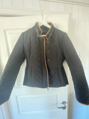 Svart quiltad jacka från Zara Woman - Säljer en svart quiltad jacka från Zara Woman med orange kantband och snygga detaljer vid kragen. Jackan har figurnära passform, två fickor och knäppning framtill. Perfekt för dig som vill ha en stilren och trendig look.