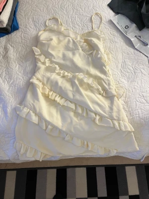 Krämvit volangklänning med smala band - Söt kort klänning i krämvit färg med smala axelband och omlott-look. Klänningen har flera diagonala volangdetaljer och är tillverkad i ett mjukt, lätt tyg som passar perfekt till varma dagar.