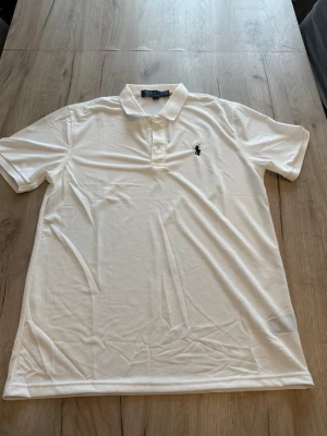 Vit piké från Polo Ralph Lauren - Klassisk vit pikétröja från Polo Ralph Lauren med den ikoniska loggan broderad på bröstet. Tröjan har korta ärmar, krage och knappar framtill. Tillverkad i mjuk bomull som känns skön mot huden.