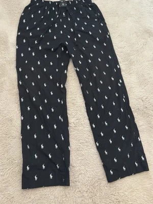 Svarta pyjamasbyxor Polo Ralph Lauren - Snygga svarta pyjamasbyxor från Polo Ralph Lauren med vitt logomönster över hela byxan. Byxorna har resår i midjan och knappgylf för extra komfort. Perfekta för chill hemma eller mysiga kvällar.