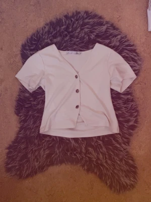 Vit kortärmad blus med knappar - Stilren vit blus med v-ringning och korta ärmar. Toppen har tre dekorativa knappar framtill och är lätt croppad i modellen. Perfekt att matcha med både jeans och kjol för en trendig look.