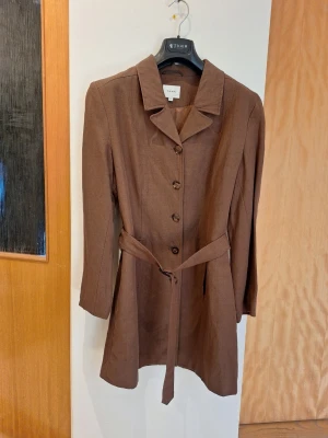 Brun kavaj i med bälte från Lindex - Fodrad brun kavaj i linnemix från Lindex med klassisk knäppning framtill och matchande midjebälte. Lång ärm och rak passform ger en clean och stilren look. 