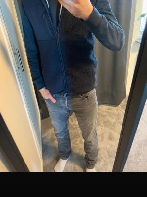 Mörkblå dunväst med dragkedja - Snygg mörkblå dunväst med dragkedja framtill och hög krage. Västen har en mix av slät och fleece-liknande yta som ger en cool kontrast. Perfekt att slänga över en hoodie för extra värme. Enkel och stilren design utan synliga loggor.